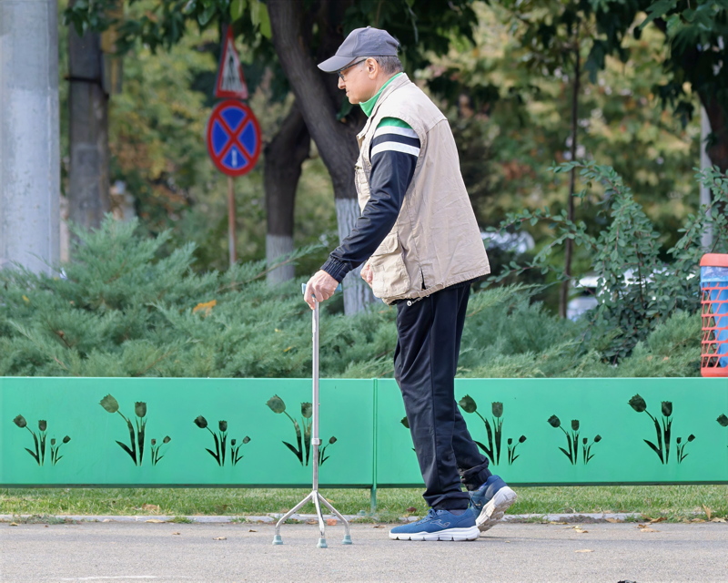29 - An older man wearing a cap, a beige vest, and dark pants as he walks outdoors on a paved path using a multi-pronged quad cane for support
